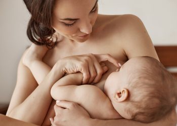 Young tender nude mother breastfeeding hugging her newborn baby sitting in bed at morning. Copy space.